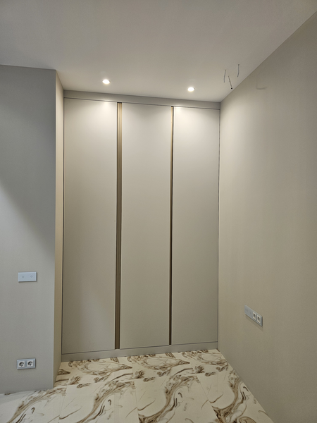 Корпусный шкаф с распашными дверями в нише помещения, выполненный в спокойной нейтральной цветовой гамме. Фасады разделены вертикальными декоративными элементами, создающими ритм и визуальную стройность конструкции. Встроенная мебель аккуратно вписана между стенами, с точной подгонкой по размерам и продуманным расположением светильников для равномерного освещения зоны хранения.