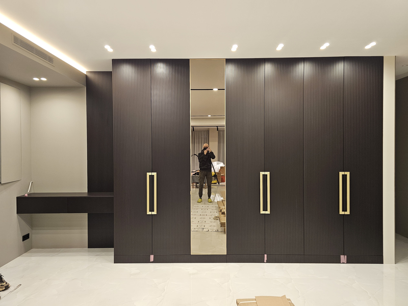Корпусный шкаф во всю стену с вертикально рифлёными фасадами тёмного оттенка, интегрированными ручками в отделке под латунь и центральной зеркальной дверью в полный рост, выполненный как архитектурный элемент интерьера с точной подгонкой по потолку и полу