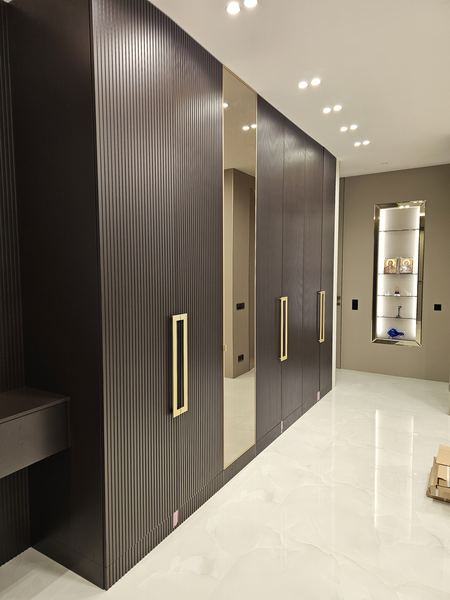 Корпусный шкаф во всю стену с рифлёными распашными фасадами, зеркальной центральной дверью и акцентными вертикальными ручками, выполненный по индивидуальному проекту с точной геометрией и встроенным освещением