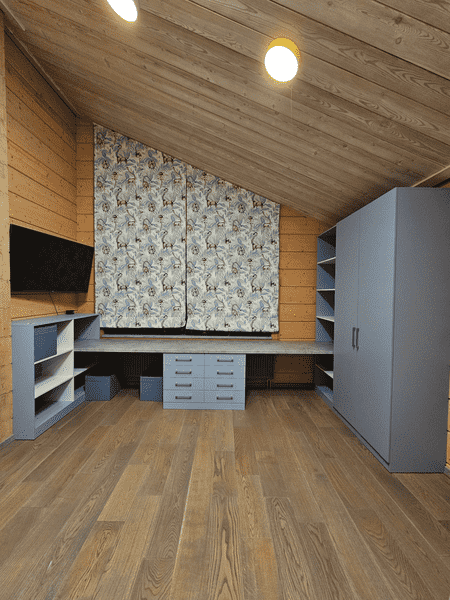 Общий вид детской комнаты на мансарде с индивидуальной мебелью: длинная рабочая столешница вдоль стены, система хранения с ящиками и шкаф для одежды, спроектированные по размерам помещения и с учётом сложной геометрии.