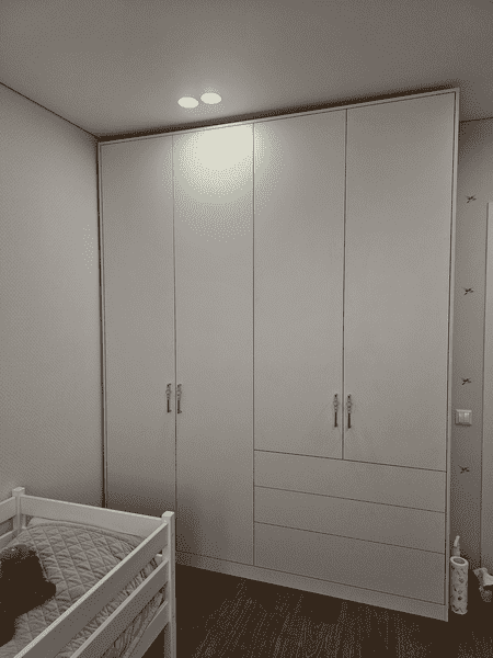 Встроенный корпусный шкаф в детской комнате с распашными фасадами во всю высоту, выполненный в белом матовом исполнении. Шкаф аккуратно вписан в нишу помещения, имеет лаконичную геометрию, скрытый цоколь и симметричную композицию. Нижний блок оснащён выдвижными ящиками для хранения детских вещей и текстиля, верхние секции предназначены для одежды на плечиках. Решение подчёркивает чистоту интерьера и визуально расширяет пространство комнаты.