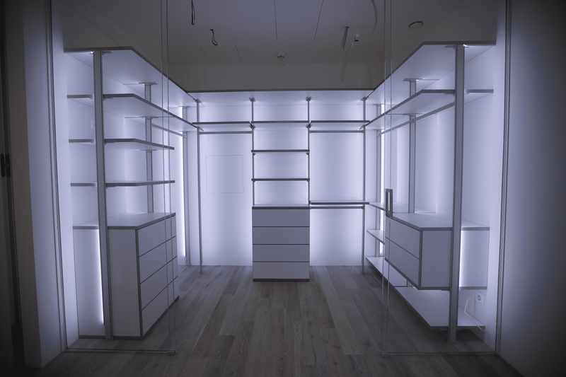 Дизайнерская открытая гардеробная с продуманной эргономикой, центральным островным комодом, симметричными секциями для хранения одежды и аксессуаров, стеклянными дверями и мягкой рассеянной подсветкой, подчёркивающей геометрию системы