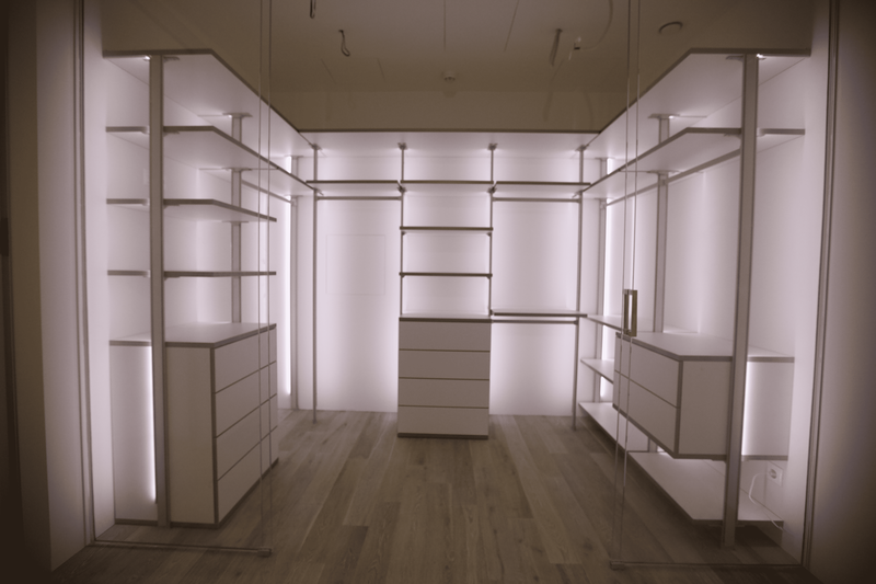 Панорамный вид гардеробной комнаты с центральной осью, открытыми стеллажами, встроенными комодами и стеклянными перегородками, демонстрирующий масштаб, продуманную эргономику и премиальный уровень исполнения