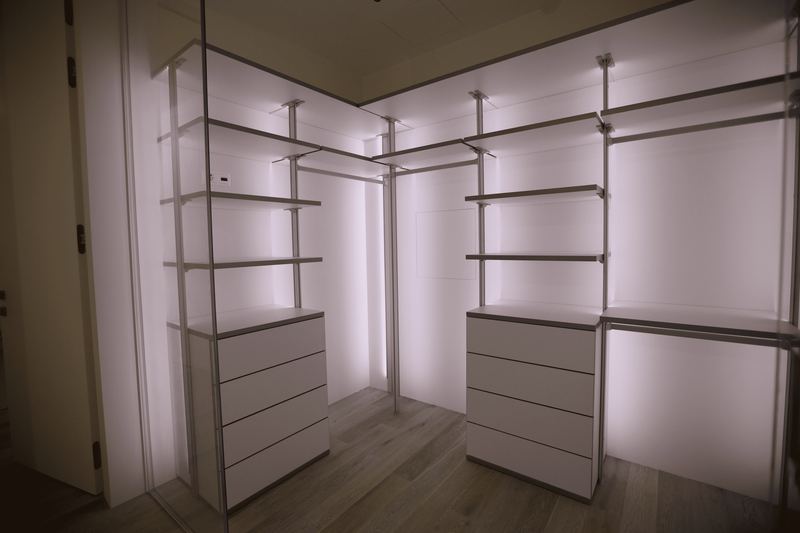 Открытая гардеробная система с металлическими стойками и полками, симметричная планировка с двумя блоками выдвижных ящиков, интегрированной вертикальной подсветкой по задней стенке и стеклянными раздвижными перегородками, создающими эффект лёгкости и архитектурной чистоты пространства