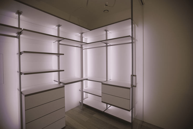 Современная открытая гардеробная комната с модульной системой хранения, металлическим каркасом, горизонтальными полками и встроенными комодами, выполненная в светлой палитре с равномерной контурной подсветкой по периметру ниш